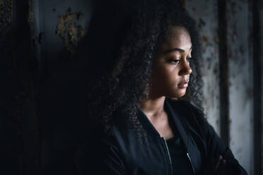 Side view portrait of sad mixed- race teenager girl standing indoors in abandoned building. - HPIF13697