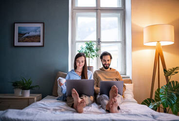 Front view of young couple using laptops on bed indoors, home office concept. - HPIF13804