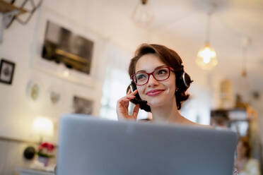 Porträt einer jungen Frau mit Headset und Laptop in einem Café, beim Arbeiten. - HPIF13861