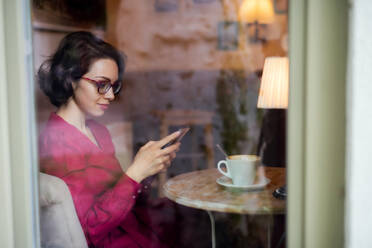 Junge Frau mit Kaffee in einem Café sitzend, Smartphone benutzend, Aufnahme durch Glas. - HPIF13862