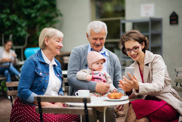 Glückliches älteres Ehepaar mit Tochter und kleiner Enkelin sitzt im Freien in einem Café und benutzt ein Smartphone. - HPIF13869