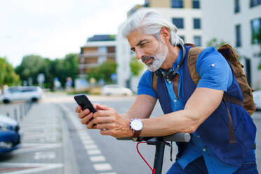 Porträt eines reifen Mannes, der mit einem Elektroroller in der Stadt unterwegs ist und ein Smartphone benutzt. - HPIF13874