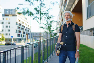 Front view portrait of mature man with skateboard outdoors in city, going back to work. - HPIF13881