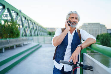 Portrait of mature man with electric scooter standing outdoors in city, using smartphone. - HPIF13885