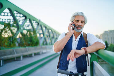 Portrait of mature man with electric scooter standing outdoors in city, using smartphone. - HPIF13886