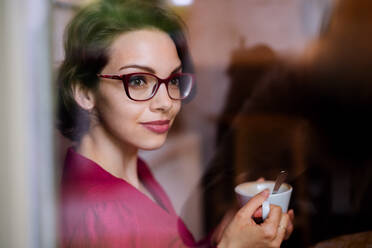 Young woman with coffee indoors in cafe. Shot through glass. Copy space. - HPIF13895