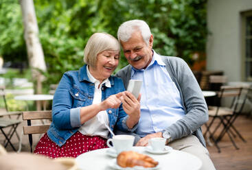 Porträt eines glücklichen, verliebten älteren Paares, das im Freien in einem Café sitzt und ein Smartphone benutzt. - HPIF13899