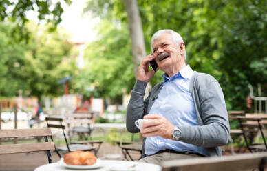 Lachender älterer Mann mit Smartphone, der im Freien in einem Café sitzt und telefoniert. - HPIF13900