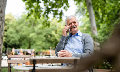 Laughing senior man with smartphone sitting outdoors in cafe, making phone call. - HPIF13901