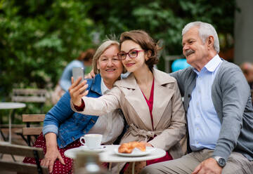 Ein junger Mann hält einen Moment mit seinen älteren Eltern in einem Straßencafé fest, indem er sein Smartphone für ein Selfie benutzt - HPIF13907