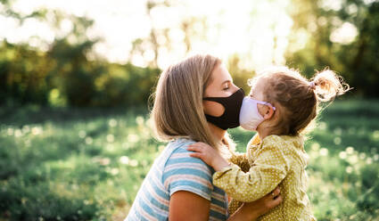 Seitenansicht einer glücklichen Mutter, die ihre kleine Tochter auf einem Ausflug in die Natur küsst und eine Gesichtsmaske trägt. - HPIF13966