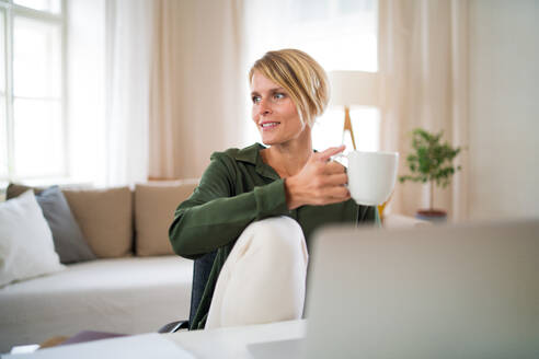 Porträt einer glücklichen Geschäftsfrau in einem Büro, die am Schreibtisch sitzt und eine Tasse Tee in der Hand hält. - HPIF14012