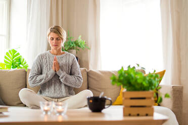 Portrait of woman indoors at home doing yoga, on sofa, mental health and meditation concept. - HPIF14020