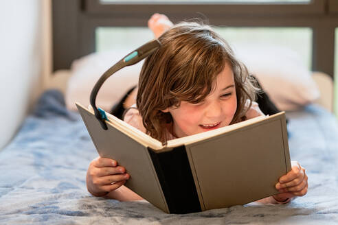 Fröhliches Mädchen mit gewelltem braunem Haar in rosa Pullover und schwarzer Hose, das auf einem weichen Bett liegt und ein interessantes Buch mit Leselicht liest, während es sich im Schlafzimmer ausruht - ADSF44072
