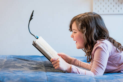 Side view of cheerful preteen girl with wavy brown hair in pink sweater and black pants lying on soft bed and reading interesting book with reading light attached while resting in bedroom - ADSF44073