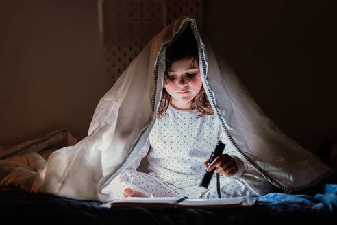 Ganzer Körper eines bezaubernden kleinen Mädchens im Pyjama, das eine Taschenlampe benutzt, während es auf dem Bett unter einer weichen Decke sitzt und ein interessantes Buch liest - ADSF44075