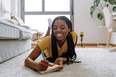 Positive junge Afroamerikanerin in lässigem T-Shirt mit langen Zöpfen, die lächelt und Ideen in ein Heft schreibt, während sie auf einem Teppich vor dem Fenster liegt - ADSF44081