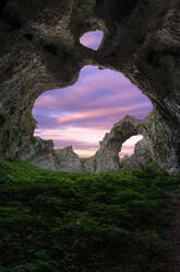 Arco de Portupekoleze Höhle mit Doppel-Loch in felsigen Formationen mit grünem Gras in malerischer Landschaft gegen bunten Sonnenuntergang Himmel in Spanien - ADSF44084