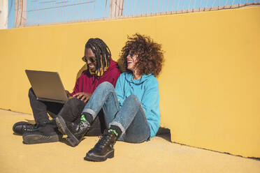 Full body of happy multiracial friends in colorful clothes sitting on sports ground with laptop while having fun together on sunny day - ADSF44105