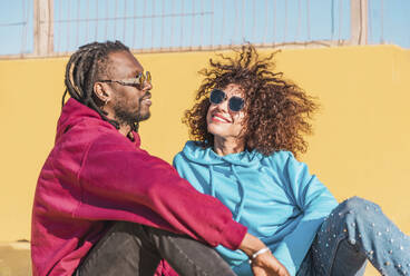 Optimistic multiracial couple in bright clothes and sunglasses laughing looking at each other sitting against yellow wall - ADSF44109