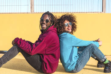 Side view of calm multiracial couple in casual wear and sunglasses sitting back to each other looking at each other on yellow ground against metal net fence - ADSF44112