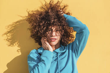 Calm curly haired female in blue hoodie touching cheek and looking at camera against yellow wall - ADSF44114