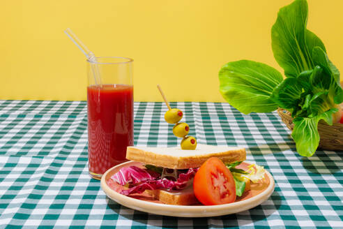 Appetitliches, gesundes Sandwich mit frischem Salat, serviert auf einem Teller mit Oliven und einer halben Tomate neben einem Glas Erfrischungsgetränk mit Glasstrohhalm vor Salatblättern auf kariertem Tischtuch - ADSF44134