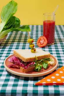 Appetitliches gesundes Sandwich mit frischem Salat, serviert auf einem Teller mit Oliven neben einem Glas Tomatensaft mit Glasstrohhalm auf einem karierten Tischtuch - ADSF44135