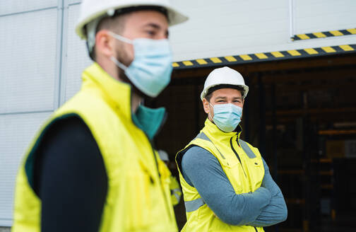 Zwei männliche Arbeiter mit Gesichtsmaske stehen vor einem Lagerhaus, Coronavirus-Konzept. - HPIF14101