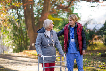 Ältere Frau mit Gehhilfe und Betreuerin oder Pflegekraft im Freien bei einem Spaziergang im Park. - HPIF14126