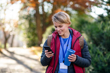 Porträt einer Pflegerin, Krankenschwester oder Mitarbeiterin im Gesundheitswesen im Freien auf dem Weg zur Arbeit, die ein Smartphone benutzt. - HPIF14129