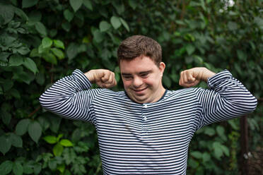 Portrait of down syndrome adult man standing outdoors at green leaves background, flexing muscles. - HPIF14262