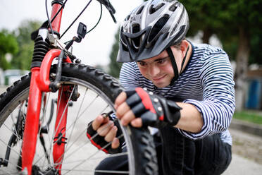 Portrait of down syndrome adult man with bicycle and helmet standing outdoors on street, repairing. - HPIF14269
