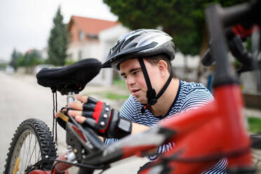 Portrait of down syndrome adult man with bicycle and helmet standing outdoors on street, repairing. - HPIF14270