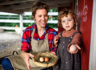 Portrait of mother with happy small daughter standing on farm, holding basket with eggs. - HPIF14499