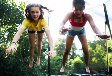 Front view of cheerful young teenager girls friends outdoors in garden, jumping on trampoline. - HPIF14642