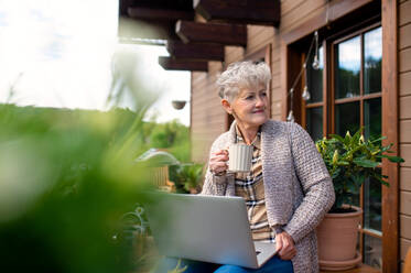 Porträt einer älteren Frau mit Laptop und Kaffee, die im Sommer auf der Terrasse sitzt und sich ausruht. - HPIF14711