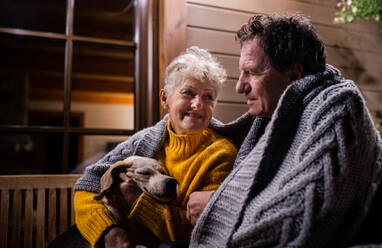 Porträt eines älteren Paares mit Hund, das abends auf der Terrasse sitzt, in eine Decke gehüllt, und sich unterhält. - HPIF14754