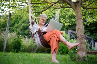 Porträt der aktiven älteren Frau mit Laptop arbeiten im Freien im Garten in der Hängematte, Home-Office-Konzept. - HPIF14806