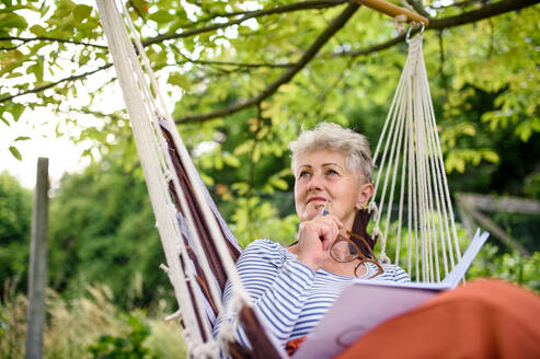 Frontansicht Porträt der glücklichen älteren Frau sitzt im Freien auf hängenden Schaukel Stuhl im Garten, entspannend. - HPIF14863