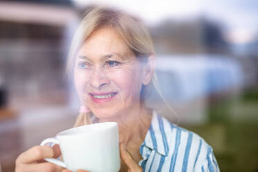Porträt einer älteren Frau mit einer Tasse Kaffee in einem Haus, aufgenommen durch ein Glas. - HPIF14918