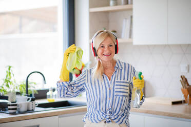 Porträt einer fröhlichen älteren Frau mit Kopfhörern und Handschuhen, die zu Hause putzt. - HPIF14926