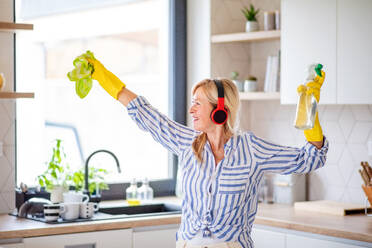 Porträt einer fröhlichen älteren Frau mit Kopfhörern und Handschuhen, die zu Hause putzt. - HPIF14927