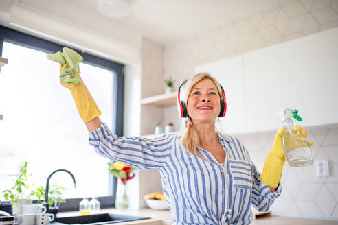 Porträt einer fröhlichen älteren Frau mit Kopfhörern und Handschuhen, die zu Hause putzt. - HPIF14930
