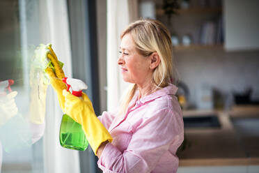 Portrait of senior woman with gloves cleaning windows indoors at home. - HPIF14947