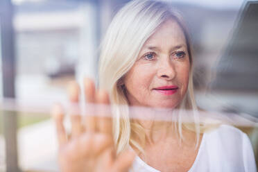 Portrait of depressed senior woman standing by window indoors at home. Shot through glass. - HPIF14964