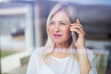 Porträt einer älteren Frau, die zu Hause steht und ihr Smartphone benutzt, aufgenommen durch ein Glas. - HPIF14967