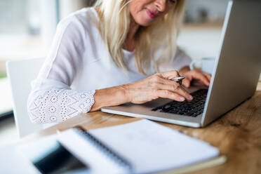 Unerkennbare ältere blonde Frau mit Laptop drinnen im Heimbüro bei der Arbeit. - HPIF14971