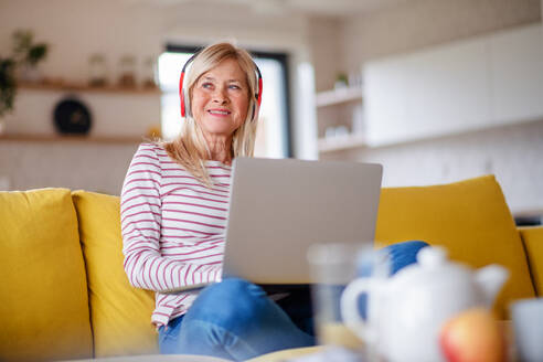 Ältere Frau mit Kopfhörern und Laptop sitzt auf dem Sofa zu Hause und entspannt sich. - HPIF14980