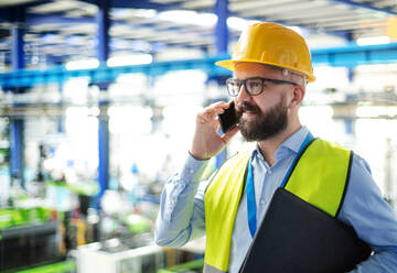 Seitenansicht eines Technikers oder Ingenieurs mit Schutzhelm in einer Industriehalle, der ein Telefon benutzt. - HPIF14994